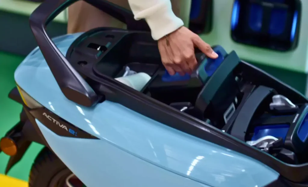 A close-up of a person's hand inserting a blue swappable battery into the compartment of a light blue Honda Activa e electric scooter, with "ACTIVA e" visible on the side.