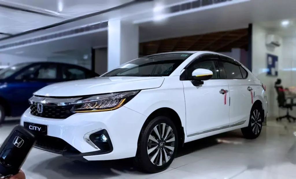 शोरूम में खड़ी नई सफेद रंग की 2025 Honda City फेसलिफ्ट कार, जिसमें शार्प LED हेडलाइट्स, डुअल-टोन अलॉय व्हील्स और साइड मिरर पर येलो एक्सेंट देखा जा सकता है। सामने हाथ में कार की चाबी भी नजर आ रही है, जो Honda के लोगो के साथ है। पीछे नीले रंग की दूसरी कार भी हल्की दिखाई दे रही है।