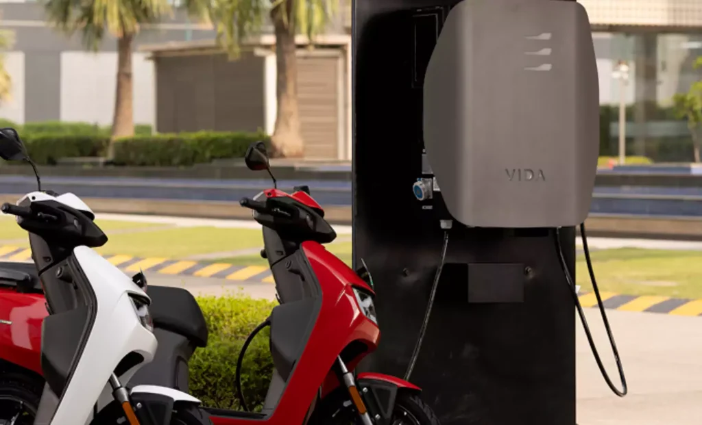 Hero Vida VX2 scooter being charged at a charging station, with a white scooter parked nearby.
