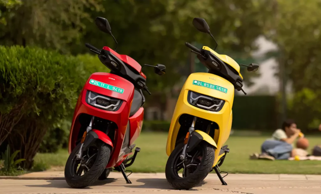 Two Hero Vida VX2 electric scooters – one red and one yellow – parked in a park, with people enjoying a picnic in the background.
